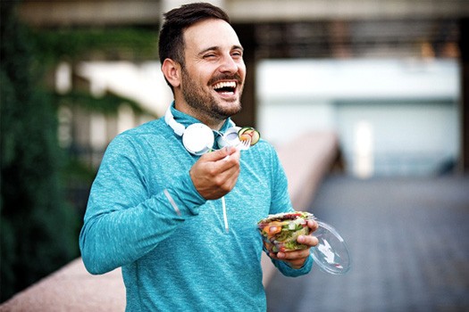 Man in active clothes outside with headphones around neck eating salad